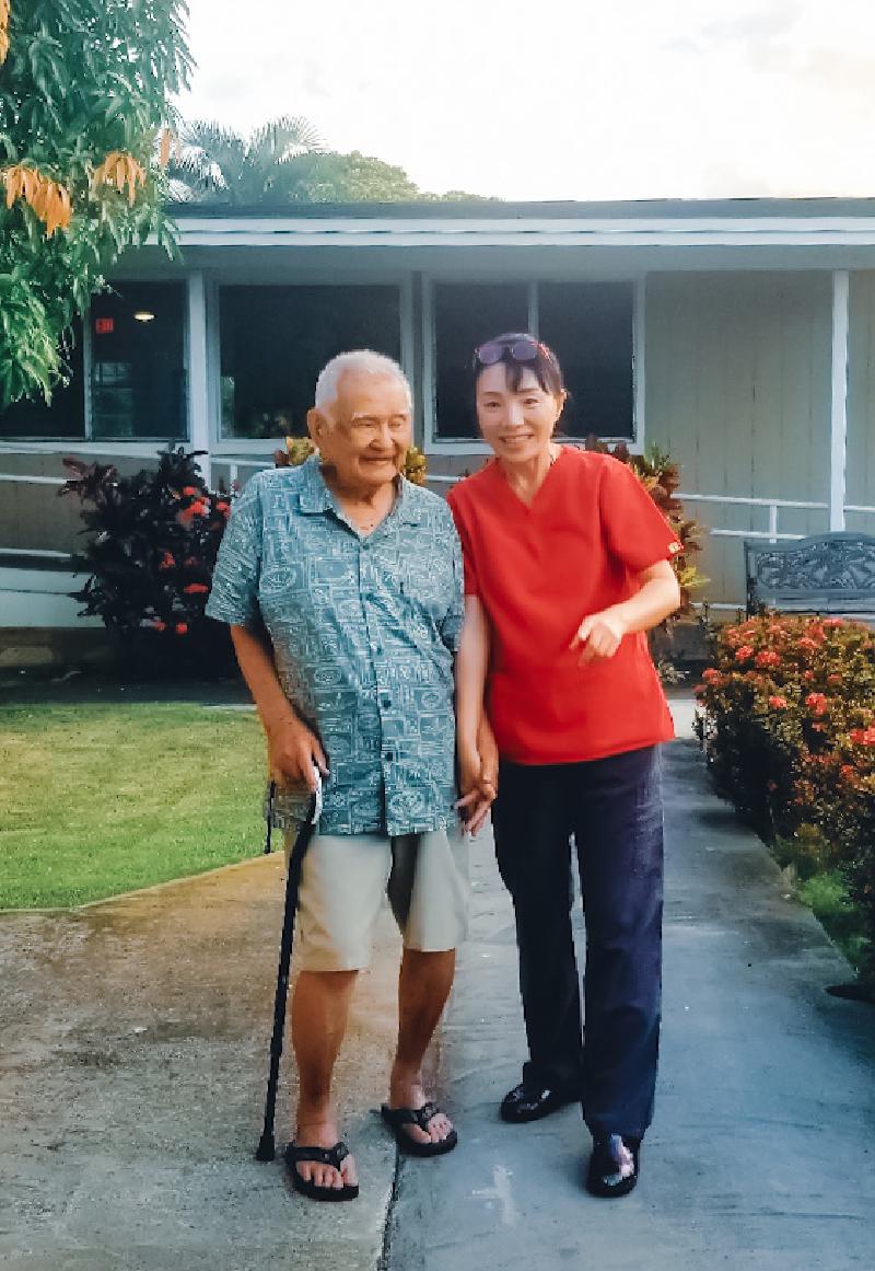 Caregiver in red scrubs walking with an elderly resident outside the facility